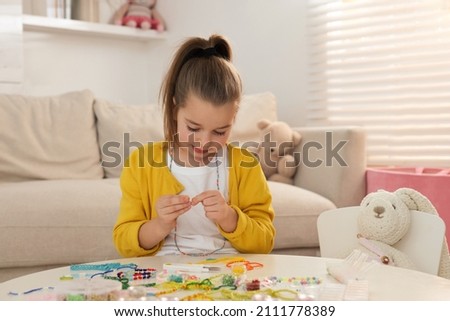 Cute little girl making beaded jewelry at table in room Royalty-Free Stock Photo #2111778389