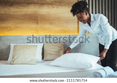 Maid working at a hotel making the bed and pillows. Maid making bed in hotel room. Housekeeper Making Bed. Young maid making bed in light hotel room. Royalty-Free Stock Photo #2111785754