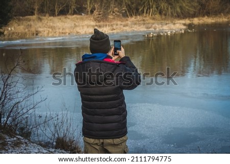 Man making photo of ducks outdoors winter rural landscape  standing on local road. fisherman guy holding mobile phone posing in village countryside. Animals wildlife organic food ecology