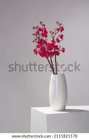 Subject shot of a white ceramic vase with branches of a thrifty wild apple-tree. The vase with the bunch of hotpink flowers is located on the white table on the gray background. Royalty-Free Stock Photo #2111821178