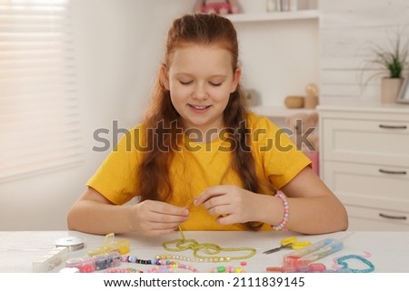 Cute girl making beaded jewelry at table in room