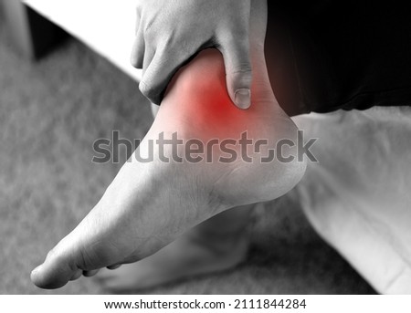  A person holding Achilles tendon by hands, suffering with pain in red spot area. Sprain ligament or Achilles tendonitis symptoms. Image in black and white with red highlights, Health care concept Royalty-Free Stock Photo #2111844284