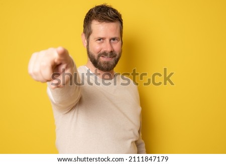 Smiling bearded man in casual sweater pointing finger at camera isolated on a yellow background