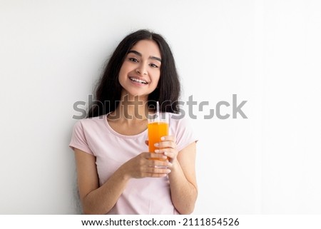 Cheerful young Indian woman holding glass of fresh orange or mango juice on white background, copy space. Happy millennial lady enjoying delicious fruit drink. Detox and healthy nutrition concept Royalty-Free Stock Photo #2111854526