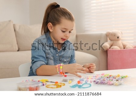 Cute little girl making beaded jewelry at table in room Royalty-Free Stock Photo #2111876684
