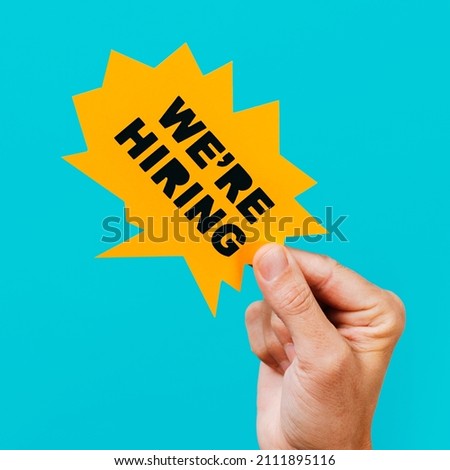 closeup of a man holding a yellow paper speech bubble with the text we are hiring in it, on a blue background, in a square format