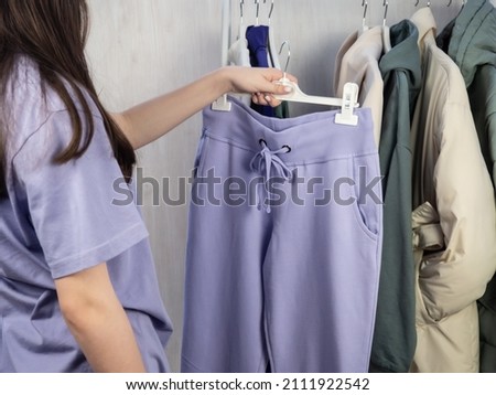 A teenage girl in a lilac T-shirt chooses what to wear. Lilac sweatpants, a white jacket and a sweatshirt hang on a hanger. Women's, youth clothes. Royalty-Free Stock Photo #2111922542