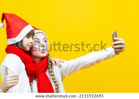 Festive Holidays. Portrait of Two Funny Caucasian Sisters Girfriends With Smartphone Taking Selfie Pictures in Santa Hat Over Yellow Background. Horizontal Image Composition