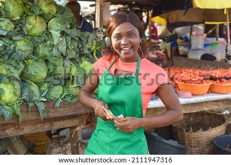 Happy African business woman or female trader wearing a green apron and standing at her vegetable stall in a market Royalty-Free Stock Photo #2111947316