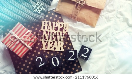 Happy New Year theme wooden letters HAPPY NEW YEAR and black painted wooden dice with number 2022 on the bed with white blanket, gift box, wooden snowflake ornaments, paper gift bag, knitted scarf.