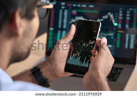 Male trader investor broker crypto analyst holding smartphone in hand analyzing stock market trading charts indexes data checking price using mobile stockmarket exchange app, over shoulder view. Royalty-Free Stock Photo #2111976929