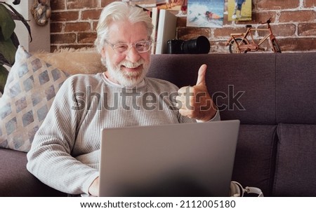Happy adult mature white haired retired smiling senior man sitting on sofa at home making distant video call with laptop computer.  Older generation and modern tech application easy usage concept.