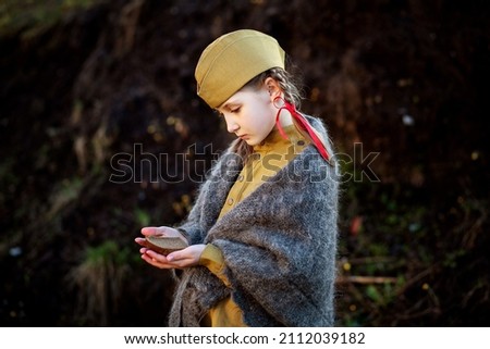 Portrait of a beautiful little girl in a military uniform rolled up in a warm shawl. She is holding a black crust of bread in her hands. Leningrad blockade. May 9 holiday. Victory Day  Royalty-Free Stock Photo #2112039182