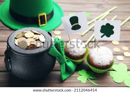 Pot of coins, green hat, bow tie, paper straws, clover leafs and cupcakes on brown wooden background