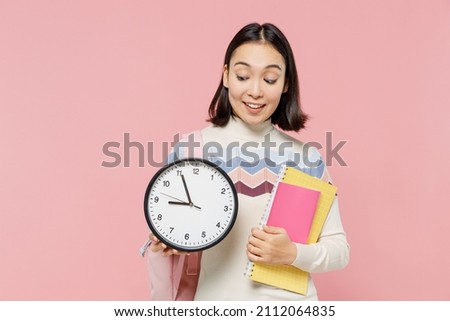 Smiling happy teen student girl of Asian ethnicity wear sweater backpack hold books look at clock isolated on pastel plain light pink background Education in high school university college concept.