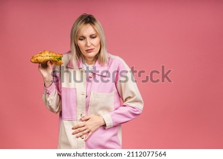 Young beautiful woman having abdominal pain isolated over pink background, holding junk food, dieting concept. 