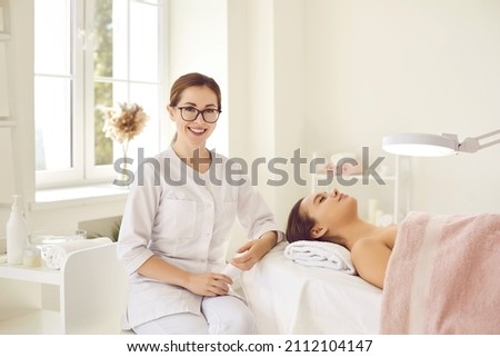 Portrait of happy dermatologist, skin therapist, beautician and skincare professional in workplace. Smiling beautiful young woman in white uniform and glasses sitting next to client on procedure table Royalty-Free Stock Photo #2112104147
