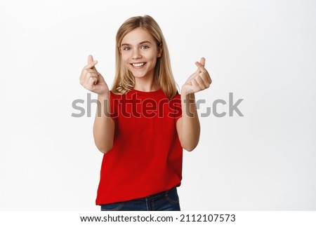 Adorable blond little girl kid, showing finger hearts and smiling, I like you gesture, standing cute in red t-shirt over white background