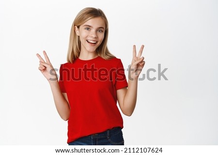 Happy, positive little girl, kid smiling and showing peace, v-sign gesture, laughing joyful, standing in red t-shirt over white background