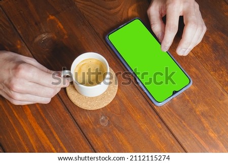 Mockup image of hands holding mobile phone with blank green screen with coffee cup on wooden table. Top view