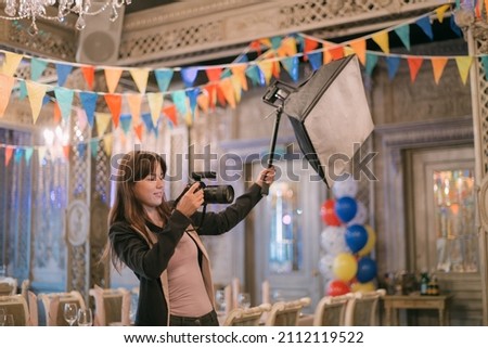 Girl photographer with a camera at work on the set in the banquet hall. A young beautiful woman - a professional photographer with a camera and a flash takes pictures on a holiday in a decorated hall Royalty-Free Stock Photo #2112119522