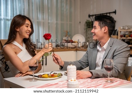 Asian young man surprise and give rose flower to beautiful girlfriend. Attractive romantic new marriage couple man and woman having dinner together to celebrate anniversary and valentines day in house Royalty-Free Stock Photo #2112119942
