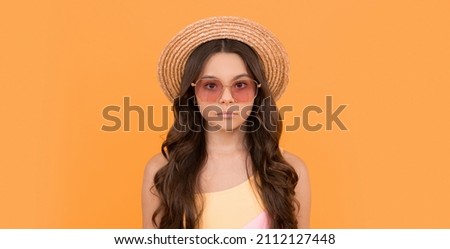serious child in summer straw hat and glasses has curly hair on orange background, fashion model