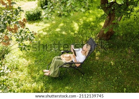 Woman reading a book in garden recliner chair. Summer vacation in the yard. Mature Scandinavian female life in retirement. Good living. View from the top. Beautiful middle aged woman resting Royalty-Free Stock Photo #2112139745