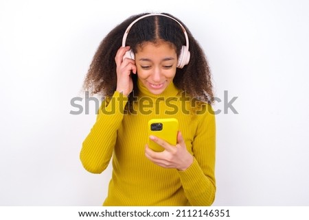 Happy beautiful teen girl with afro hairstyle wearing yellow turtleneck sweater against white background feels good while focused in screen of smartphone. People, technology, lifestyle