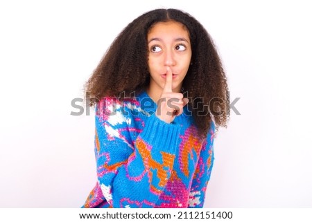 teen girl wearing colourful knitted sweater against white silence gesture keeps index finger to lips makes hush sign. Asks not to share secret