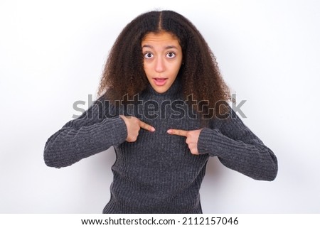 Embarrassed beautiful teen girl wearing grey turtleneck sweater against white indicates at herself with puzzled expression, being shocked to be chosen to participate in competition.