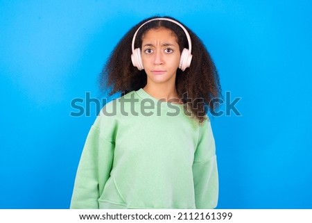 Serious displeased beautiful teen girl wearing green sweater standing against blue background looks puzzled at camera being angry wears stereo headphones listens music while walking at street