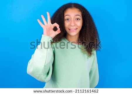 beautiful teen girl wearing green sweater standing against blue background hold hand arm okey symbol toothy approve advising novelty news