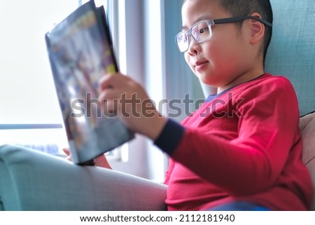 Cute boy wearing glasses, smiling and reading a fun graphic comic novel book near window at home. Child learning activity background.