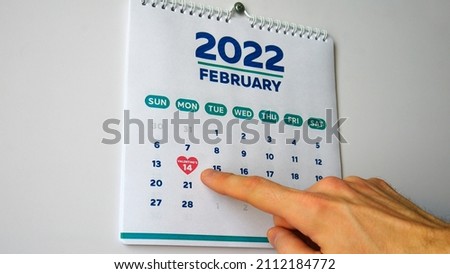 A man points finger at Valentine's date designed with a pink heart symbol on a February page of the calendar 2022