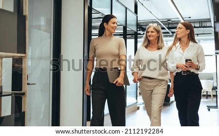 Three cheerful businesswomen walking together in an office. Diverse group of businesswomen smiling while having a discussion. Successful female colleagues collaborating on a new project. Royalty-Free Stock Photo #2112192284