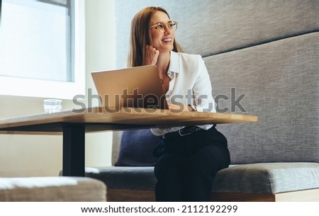 Excited businesswoman looking away thoughtfully. Happy young businesswoman smiling while contemplating business ideas. Young female entrepreneur working on a laptop in a modern workspace. Royalty-Free Stock Photo #2112192299
