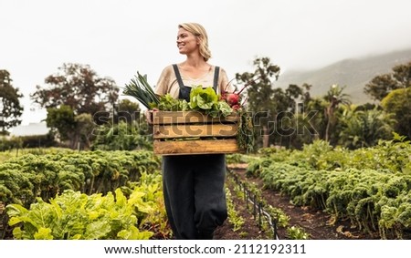 Female organic farmer gathering fresh vegetables on her farm. Happy young woman holding a box with fresh produce in her vegetable garden. Successful organic farmer smiling while harvesting. Royalty-Free Stock Photo #2112192311