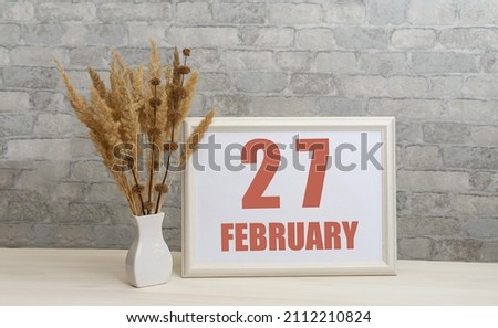 february 27. 27th day of month, calendar date.  White vase with ikebana and photo frame with numbers on desktop, opposite brick wall. Concept of day of year, time planner, winter month.