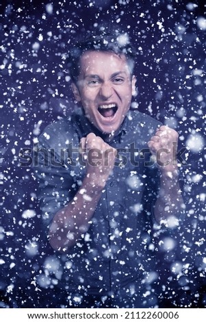 A brunette man in a shirt is joyful on a dark background. Hands into fists. Victory. Gesture of joy. Snowfall.