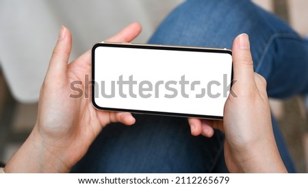 Close-up image of a female's hands holding a smartphone over her legs. Mockup of a smartphone's blank screen. Play games, watch videos.