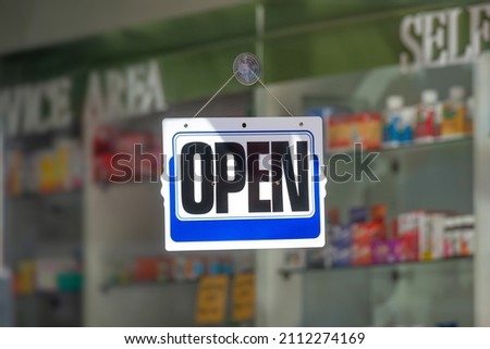 Photo of an Open sign with white and blue color over blur drug store or Pharmacy background with sunlight and shadow in the daytime. Backdrop artwork design lifestyle shop banner text concept ideas.