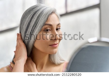 Portrait of beautyful happy middle aged mature asian woman, senior older 50s lady pampering touching gray hair looking at herself at mirror indoors. Ads of lifting anti wrinkle skin hair care spa. Royalty-Free Stock Photo #2112280520
