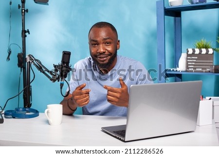 Smiling vlogger talking with audience during online live show sitting at desk with laptop and broadcasting setup. Content creator interacting with fanbase in recording studio with professional