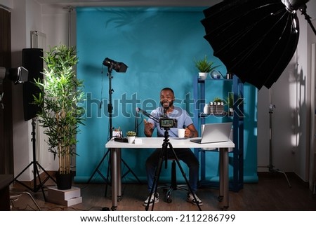 Wide view of smiling influencer interacting with audience sitting at desk holding cup in vlogging studio setup with video ligths. Content creator talking to followers in front of filming video camera.