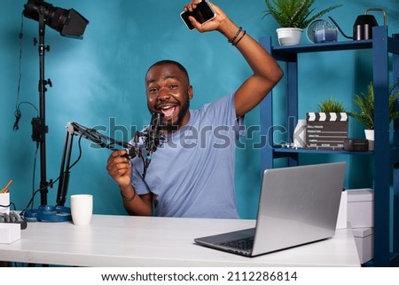 Happy vlogger raising hand in excitement holding smartphone shouting in professional microphone sitting at desk in vlogging studio. Content creator feeling joy after receiving positive feedback.
