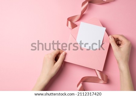 First person top view photo of valentine's day decorations female hands holding open pink envelope with paper sheet and pink silk curly ribbon on isolated pastel pink background with blank space
