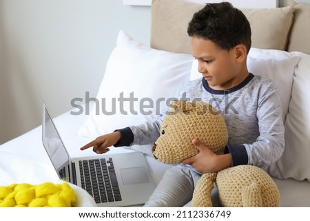 Little African-American boy with laptop and teddy bear watching cartoons on bed