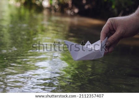 A hand lowers a white paper boat on the water. Man holds the homemade boat. Shooting at water level. Selective focus. Blurred motion.