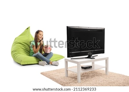 Girl sitting on a green bean bag armchair with a box of popcorn and watching tv isolated on white background